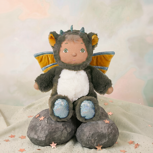 Plush toy dragon with a child-like face sitting on a rock, surrounded by star-shaped confetti. Cozy Day Dinkum Flint by Olli Ella – Magical Dragon Plush Toy Suitable for Newborns