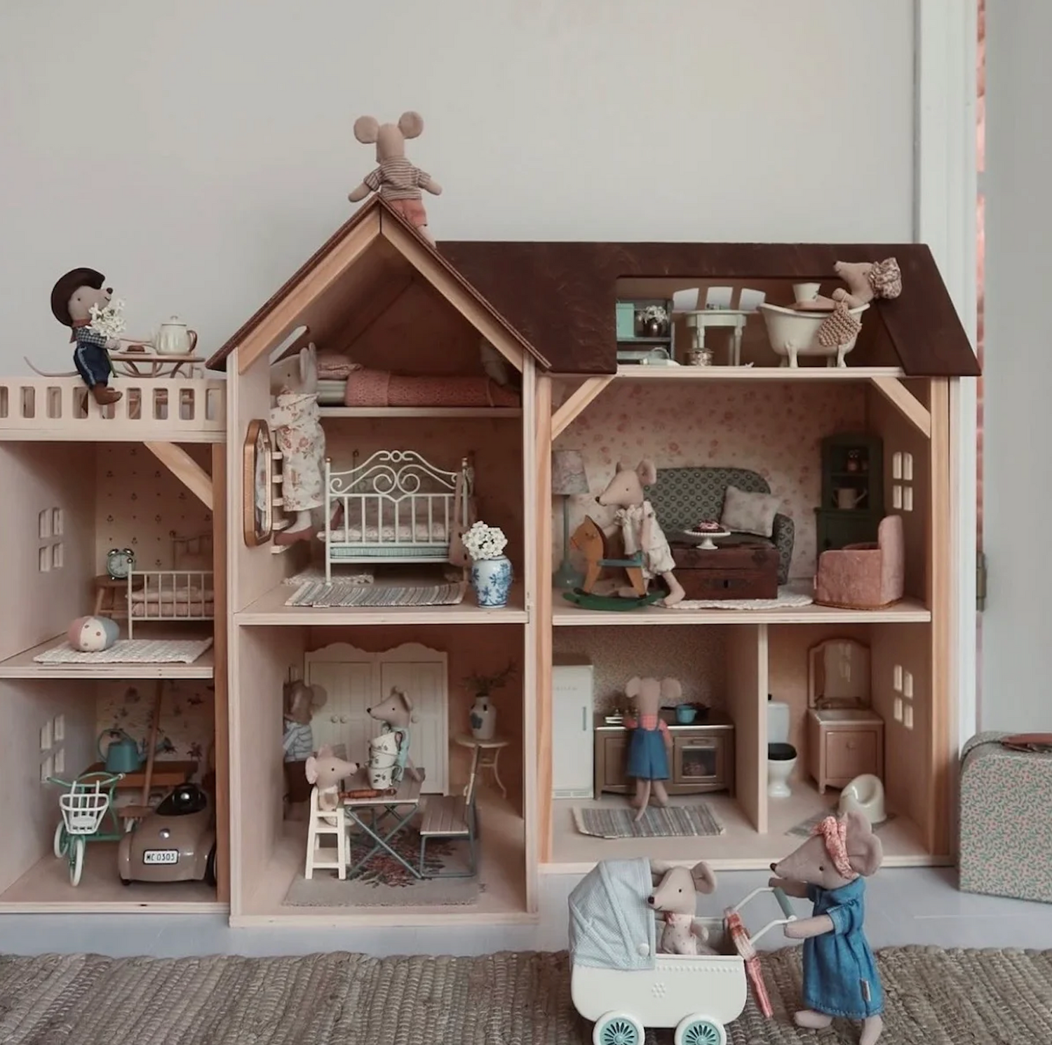 A large wooden dollhouse with multiple rooms and shelves, decorated with miniature furniture, figurines, and accessories. Several mouse-like toy figures are arranged inside and outside the dollhouse, including one pushing a stroller in the foreground.