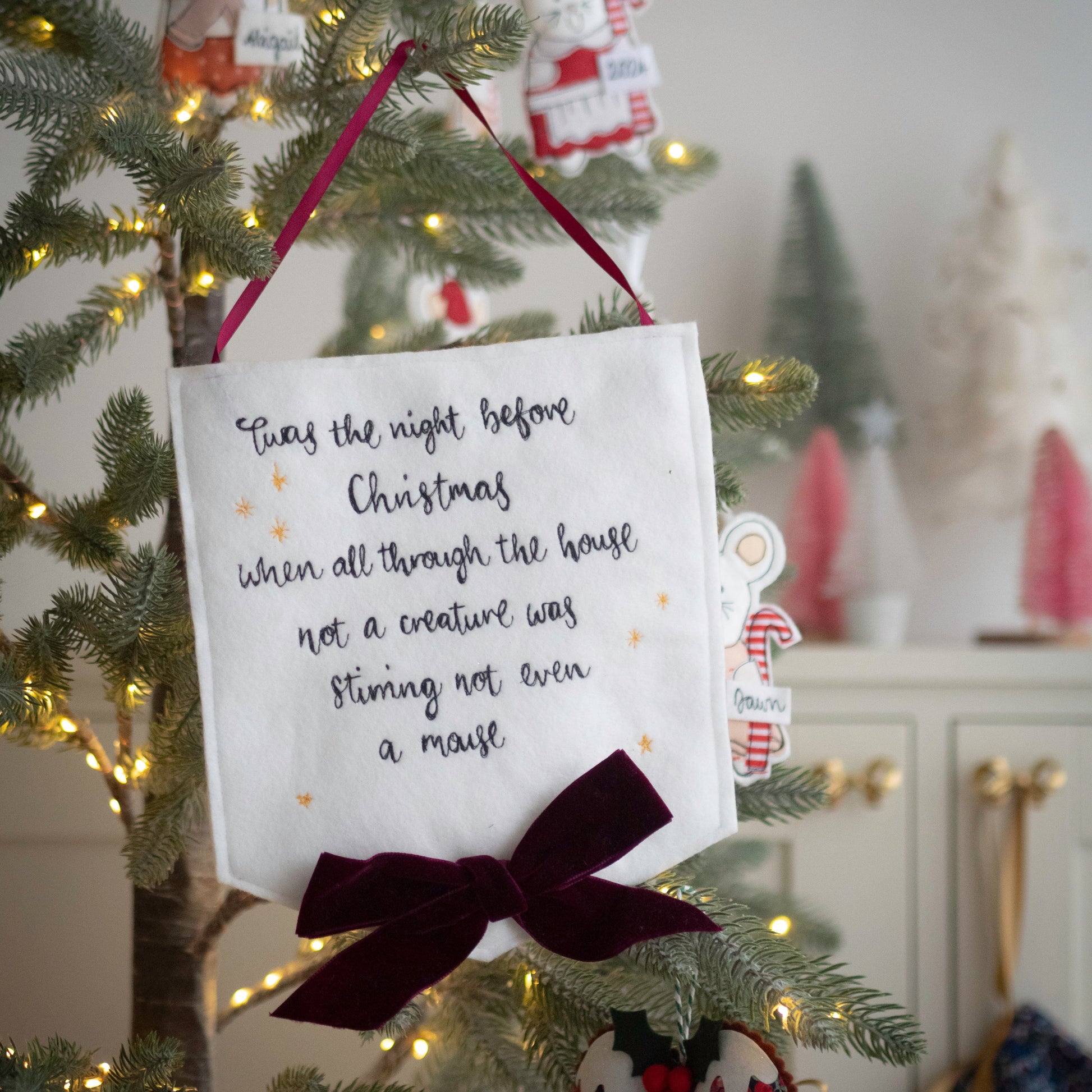 Decorative sign Christmas banner with the quote Twas the night before Christmas, a hand embroidered white fabric banner sign by Girl Friday Embroidery at Mouse in a Box with a Christmas tree in the background