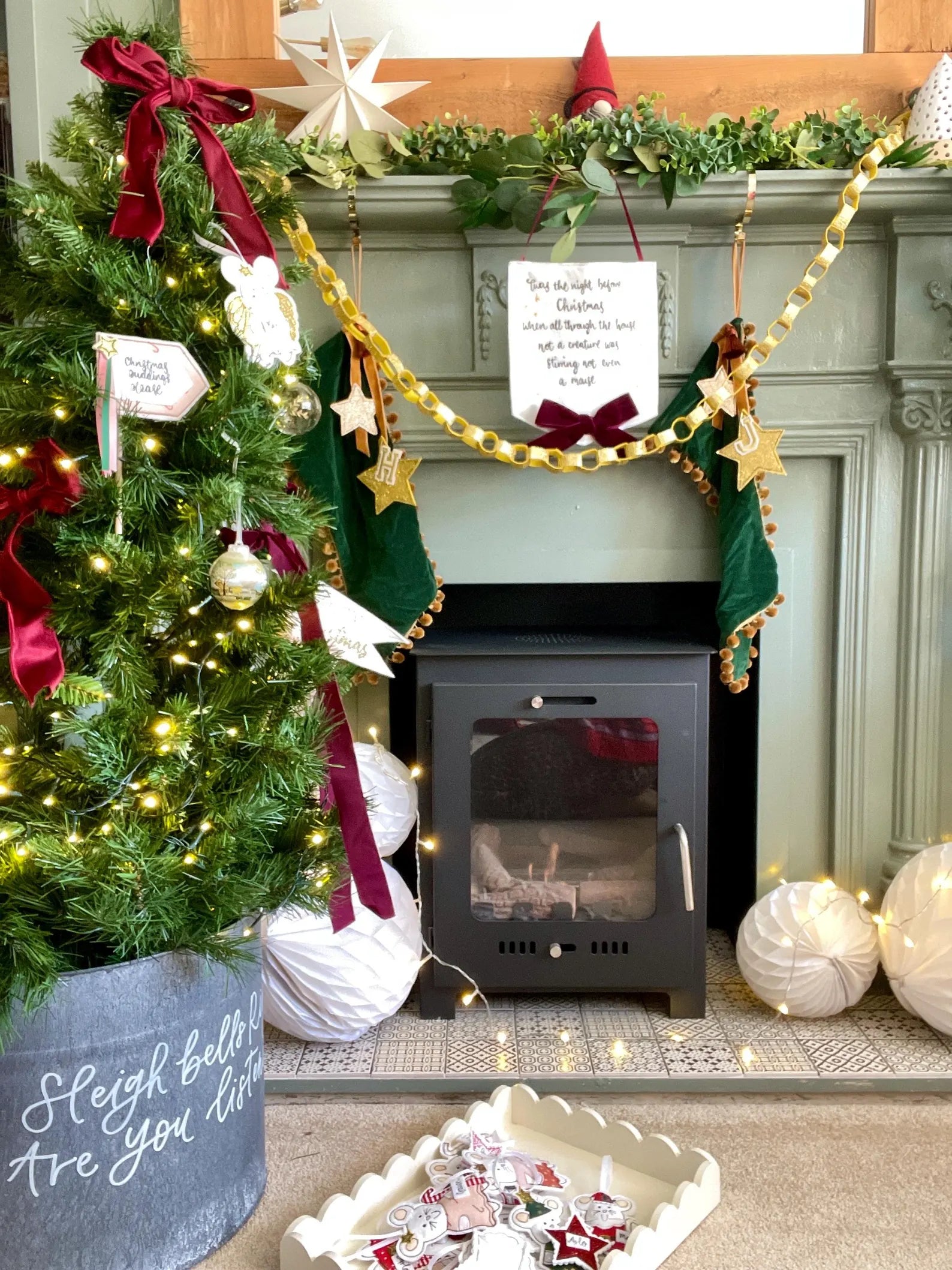 Decorated Christmas tree with lights and Christmas banner with the quote Twas the night before Christmas in front of a fireplace.