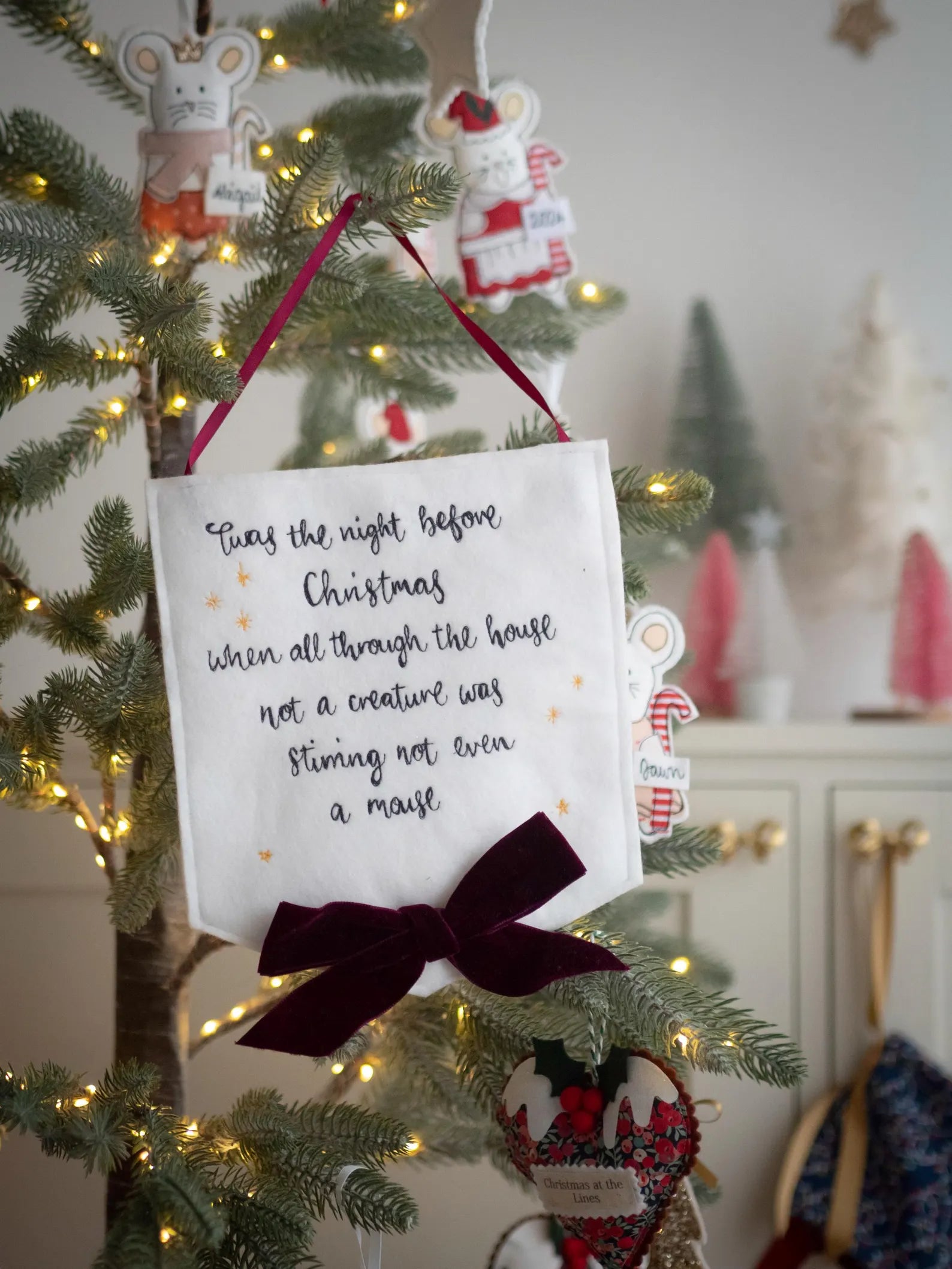 Decorative white fabric banner with the quote Twas the night before Christmas hand stitched text on a Christmas tree with lights and ornaments.